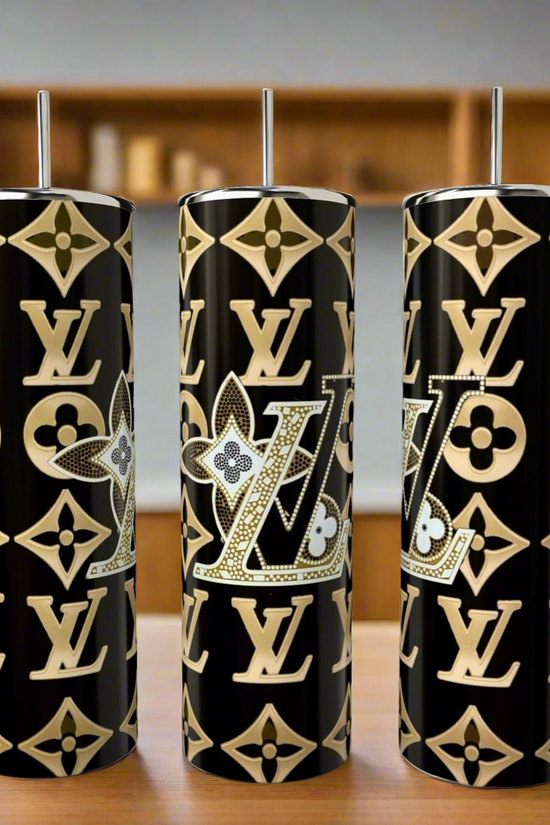 Three black Kreative Kreationz LV Glitz 20oz Skinny Tumblers with metallic lids and straws sit on a wooden counter, each adorned with a gold monogram design. These eco-friendly tumblers add style to any kitchen.