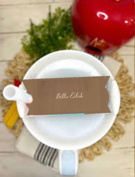 A white cup on a saucer rests on a woven mat, with a Kreative Kreationz "Billie Eilish Stanley Lid Topper" displayed nearby. In the background, green foliage and an apple-shaped lid topper create a whimsical vibe.