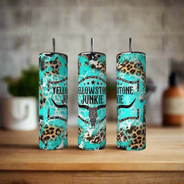 The Kreative Kreationz Teal Yellowstone Junkie 20oz Tumbler features a bull skull graphic and "Yellowstone Junkie" text, shown on a wooden surface alongside brown and leopard-print stainless steel tumblers; the background is softly blurred.