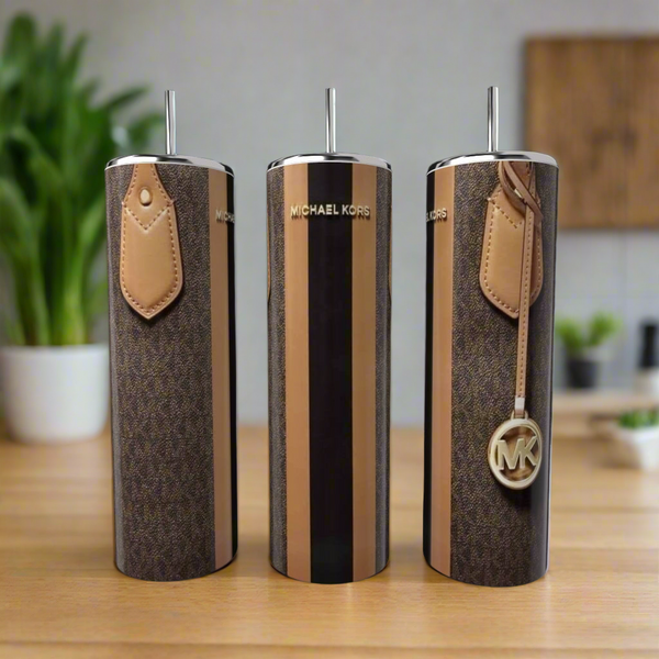 Three Kreative Kreationz MK Stripe 20oz Tumblers with straws are arranged on a wooden surface. Each features a brown and tan stripe design with logo details, and a plant plus blurred kitchen items appear in the background.