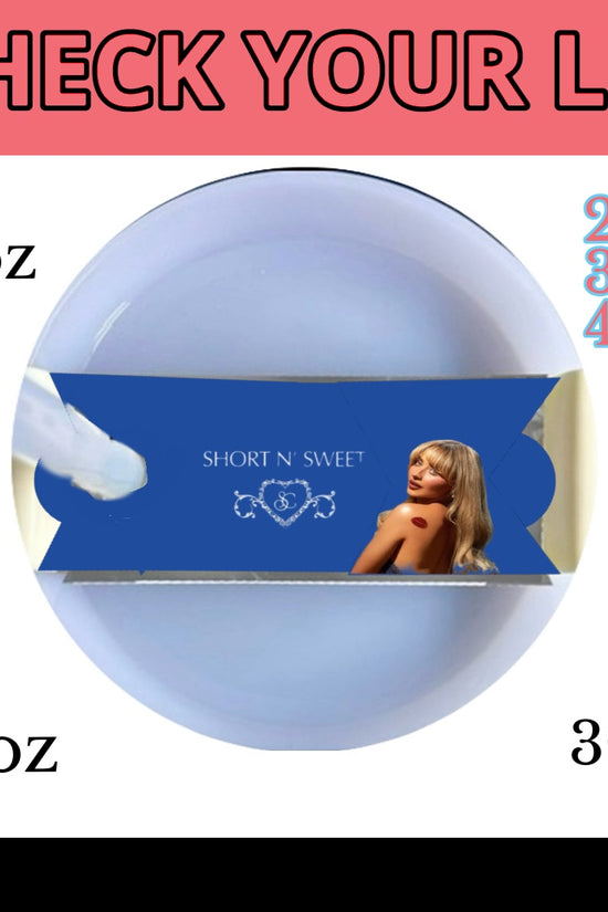 A white mug with a light blue button feature, covered by a lid. On the lid sits a blue, rectangular packaging featuring an image of a woman with blonde hair in a black outfit. The text on the packaging reads 
