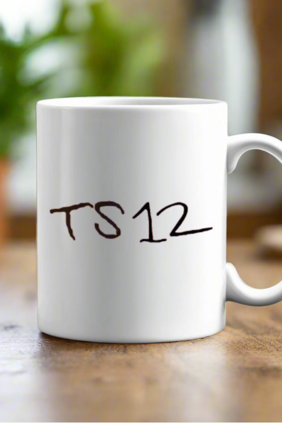 The TS12 Ceramic Mug by Kreative Kreationz rests on a wooden table, displaying the text 