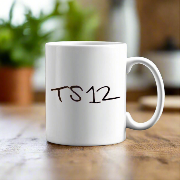 The TS12 Ceramic Mug by Kreative Kreationz rests on a wooden table, displaying the text "TS12" in black. A blurred green plant and kitchen items are seen in the background.