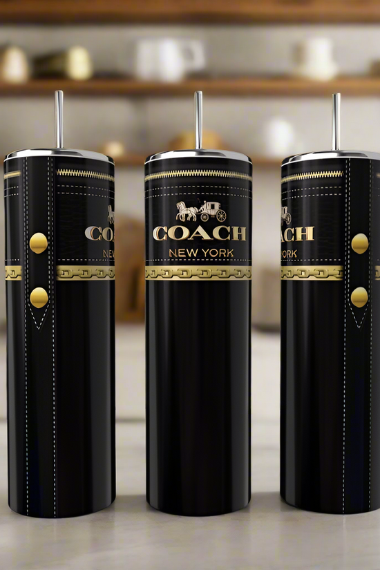 Three Coach Black & Gold 20oz Skinny Tumblers from Kreative Kreationz, each with a metal straw and gold logo, stand elegantly on a light kitchen counter with softly blurred shelves and mugs in the background.