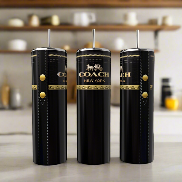 Three Coach Black & Gold 20oz Skinny Tumblers from Kreative Kreationz, each with a metal straw and gold logo, stand elegantly on a light kitchen counter with softly blurred shelves and mugs in the background.