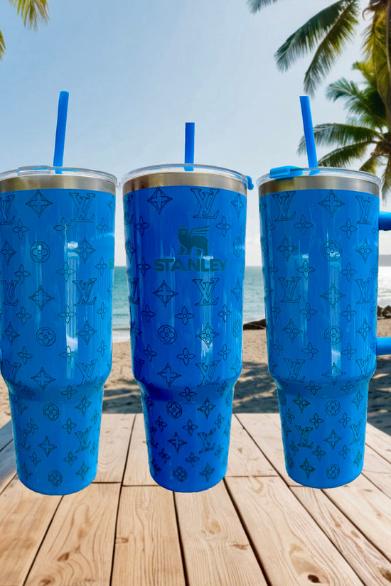 Three blue LV & Stanley Engraved 40oz Tumblers by Kreative Kreationz rest on a wooden table at a beach—palm trees and ocean behind them—making these personalized stainless steel tumblers ideal for your next getaway.