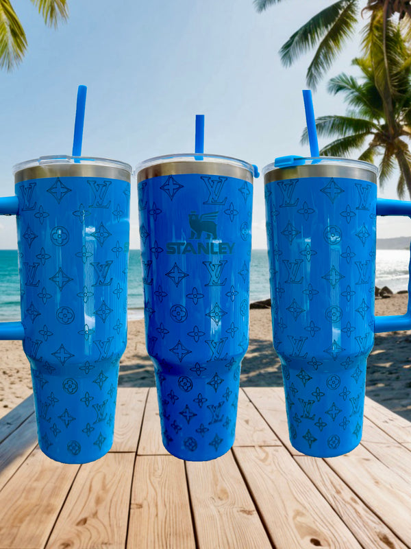 Three blue LV & Stanley Engraved 40oz Tumblers by Kreative Kreationz rest on a wooden table at a beach—palm trees and ocean behind them—making these personalized stainless steel tumblers ideal for your next getaway.