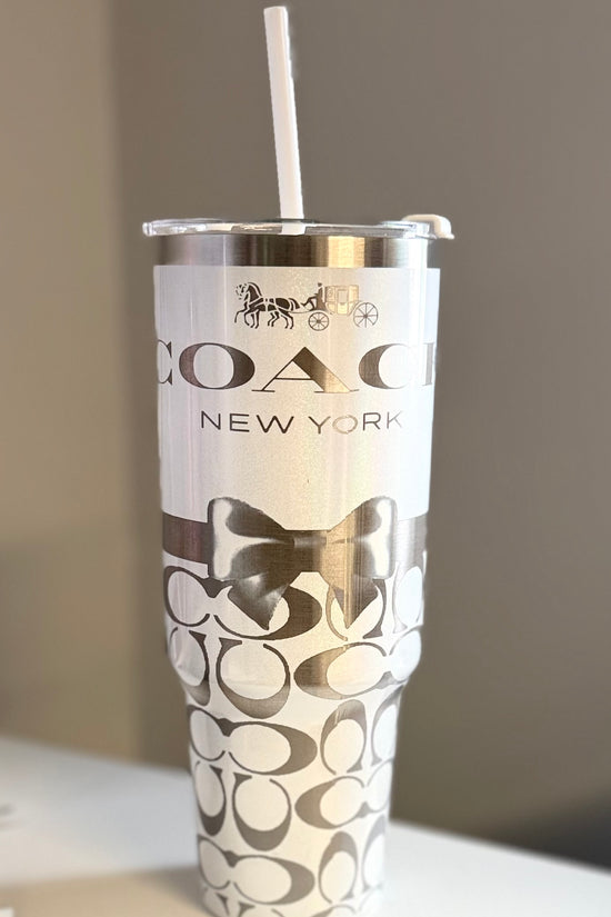 The Coach White 40oz Engraved Tumbler by Kreative Kreationz features a white straw, silver horse and carriage logo, black bow design, and overlapping 