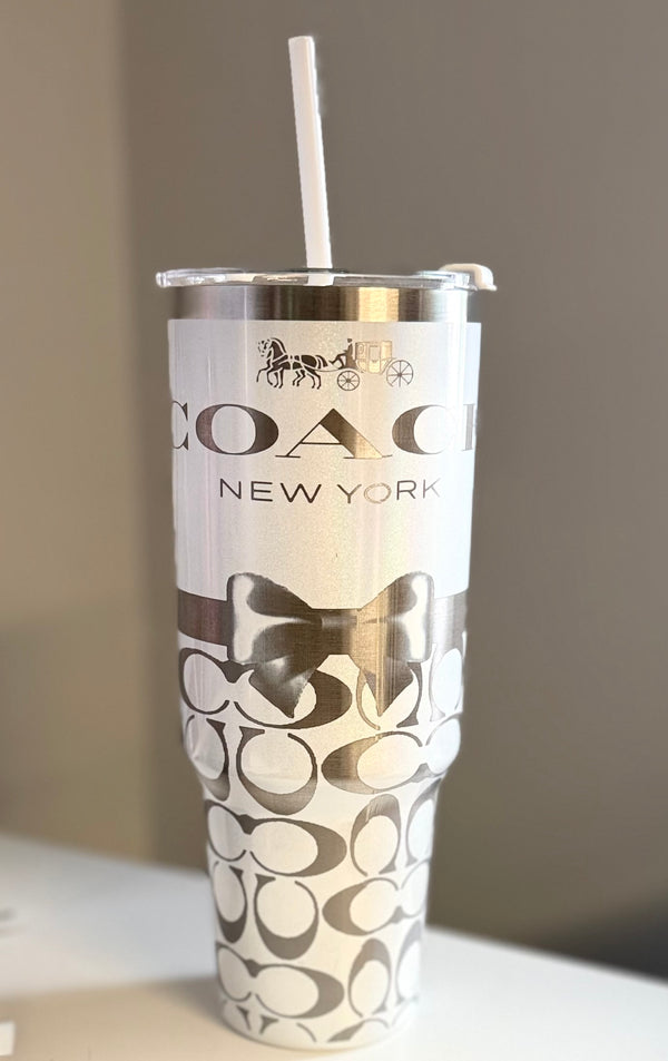 The Coach White 40oz Engraved Tumbler by Kreative Kreationz features a white straw, silver horse and carriage logo, black bow design, and overlapping "C" pattern on a white stainless steel background.
