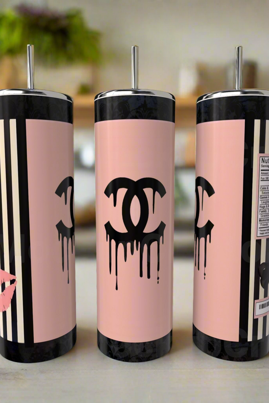 Three Kreative Kreationz Chanel 20oz Skinny Tumblers, pink with black dripping logos, stripes, and metal straws rest on a counter; one shows a kiss mark and another a playful nutrition label for eco-friendly drinkware style.