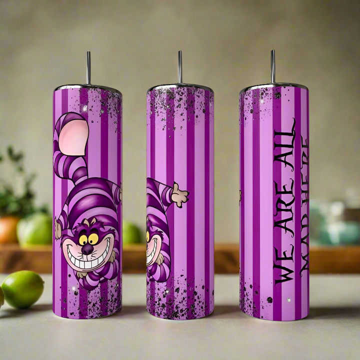 Three tall, cylindrical "We are all a Little Mad Tumbler" by Kreative Kreationz feature purple and pink stripes, the Cheshire Cat from "Alice in Wonderland," and the phrase "We are all mad here" in black text. Kitchen items blur in the background.