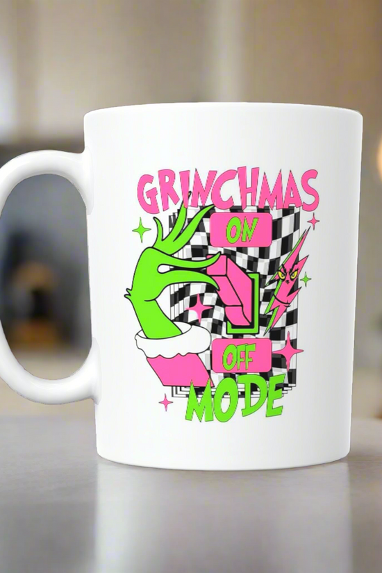 The Grinchmas Ceramic Mug by Kreative Kreationz features a green hand flipping a switch with “GRINCHMAS ON OFF MODE” in pink and green on a black-and-white checkered background—perfect for celebrating Grinchmas mode!.