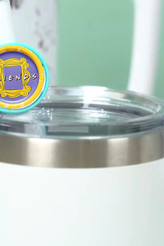 On the table is a white tumbler with a clear lid and stainless steel rim, accompanied by a circular cap featuring a yellow 