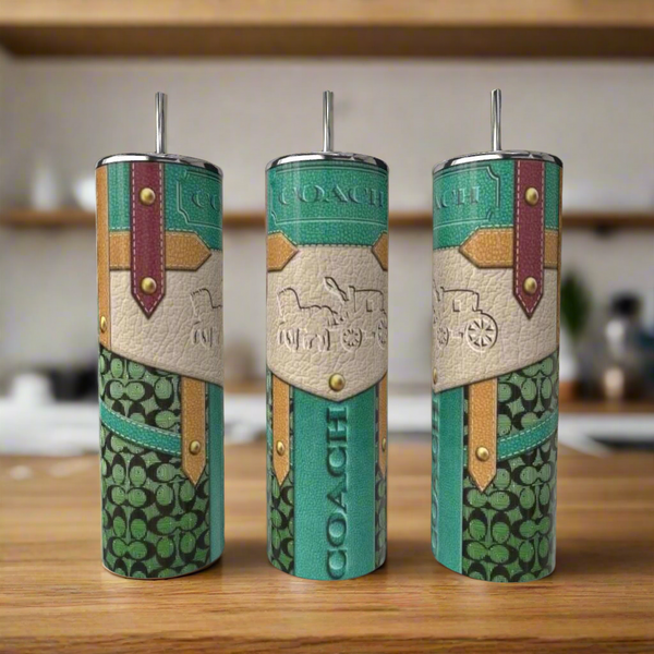 Three Coach Strap 20oz Tumblers by Kreative Kreationz, each with green, cream, and brown patchwork designs and embossed carriage logos, come with metal straws and sit on a wooden counter. A blurred kitchen is in the background.