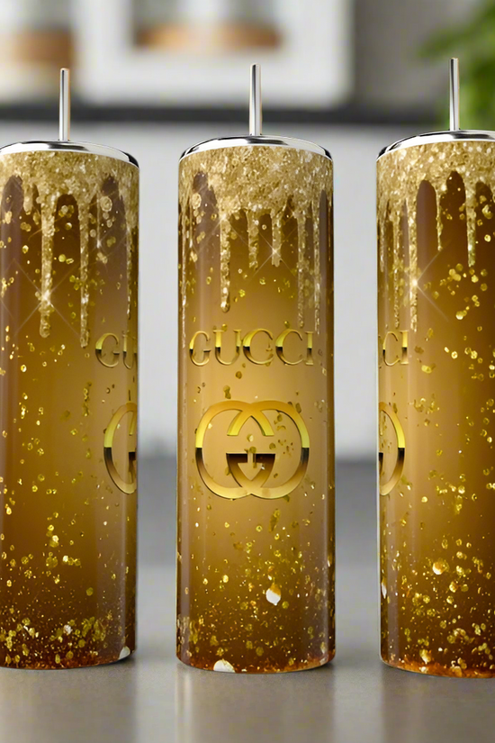 Three Gucci Gold 20oz Skinny Tumblers by Kreative Kreationz stand tall on a countertop. Filled with golden glitter and displaying the Gucci logo, they are set against a blurred backdrop of lush foliage, reflecting the sophistication of an elegant design.
