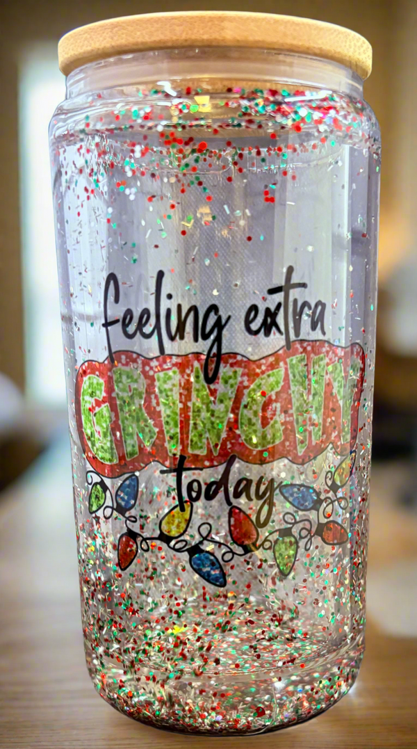 The Grinch Snowglobe Tumbler by Kreative Kreationz is a transparent 16oz jar with a bamboo lid, filled with red, green, and white glitter. It features the phrase "feeling extra GRINCHY today" and colorful Christmas lights—ideal for those who enjoy fun holiday gifts.