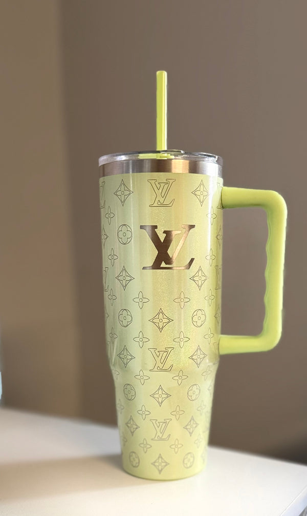 The LV Engraved Shimmer Tumbler by Kreative Kreationz is a large pale yellow stainless steel tumbler with a handle and straw, featuring the Louis Vuitton logo and monogram pattern, displayed on a light surface with a neutral background.
