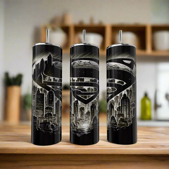 Three Kreative Kreationz Superman Modern 20oz Tumblers with black stainless steel, metallic Superman logos, and city skyline designs sit on a wooden counter with a blurred kitchen background—ideal for any fan.