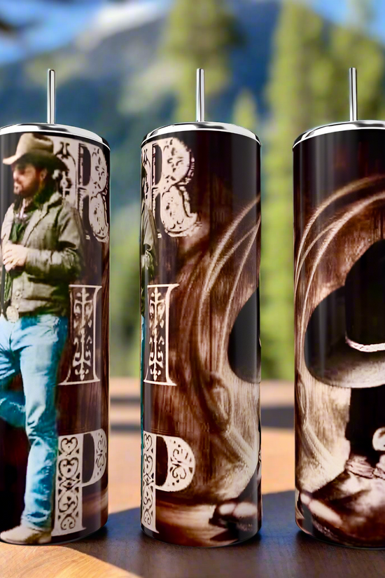 Three Kreative Kreationz Yellowstone RIP 20oz stainless steel tumblers with western-themed designs, featuring a cowboy and intricate lettering, stand proudly on a wooden surface against a blurred backdrop of forest and mountain.