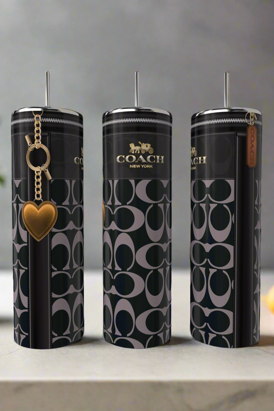 Three tall, cylindrical Coach 20oz Skinny Tumblers by Kreative Kreationz with black and gray signature patterns are on a kitchen counter; one features a gold heart keychain. A potted plant and lemons blur in the background.