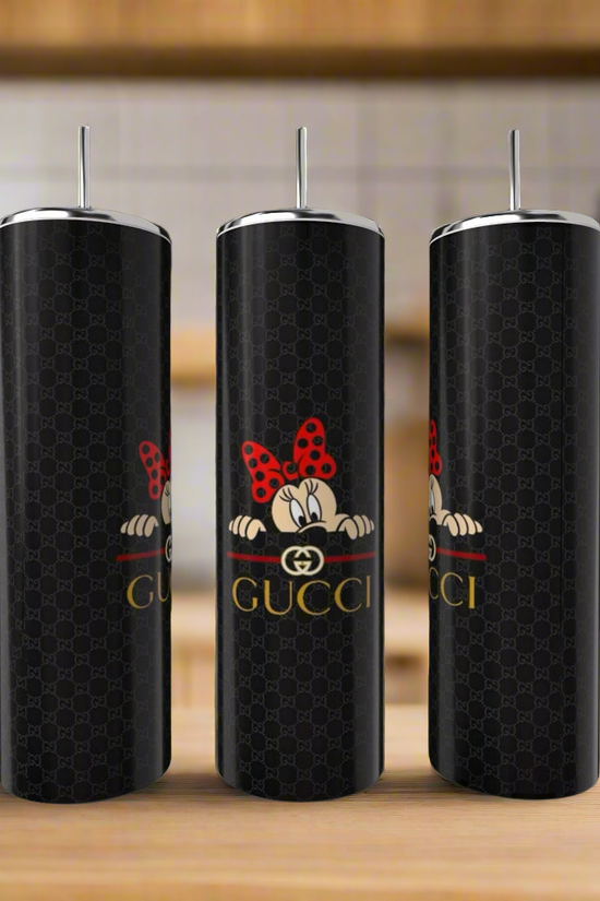 Three Gucci Mickey Mouse 20oz Skinny Tumblers by Kreative Kreationz rest elegantly on wood. Adorned with a cartoon character and red polka-dotted bow, they capture attention against a blurred kitchen backdrop dotted with plants and kitchenware.