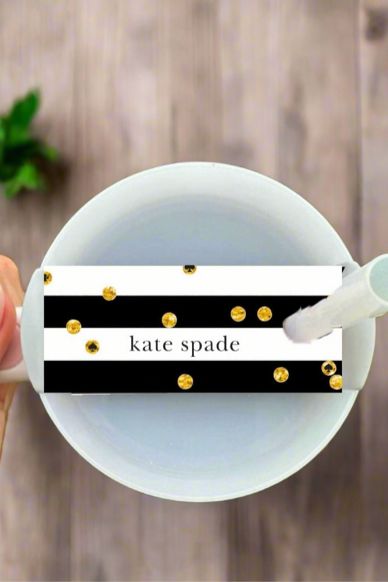 A hand holds a white mug above a wooden table, featuring the Kate Spade Stanley Lid Topper by Kreative Kreationz—a black and white striped tag with gold dots and “kate spade” text. A green plant appears in the background.