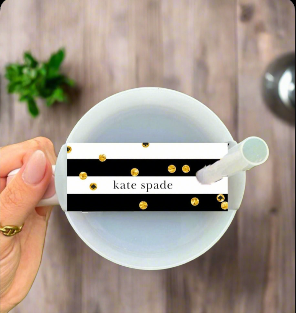 A hand holds a white mug above a wooden table, featuring the Kate Spade Stanley Lid Topper by Kreative Kreationz—a black and white striped tag with gold dots and “kate spade” text. A green plant appears in the background.