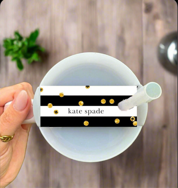 A hand holds a white mug above a wooden table, featuring the Kate Spade Stanley Lid Topper by Kreative Kreationz—a black and white striped tag with gold dots and “kate spade” text. A green plant appears in the background.