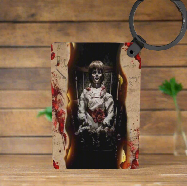 The Annabelle Inspired Keychain by Kreative Kreationz features a creepy doll in a burnt, bloodstained frame with wood and green plants in the background, offering fade-resistant colors for long-lasting impact.