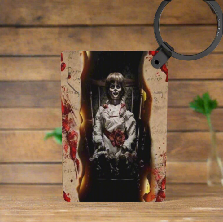 The Annabelle Inspired Keychain by Kreative Kreationz features a creepy doll in a burnt, bloodstained frame with wood and green plants in the background, offering fade-resistant colors for long-lasting impact.