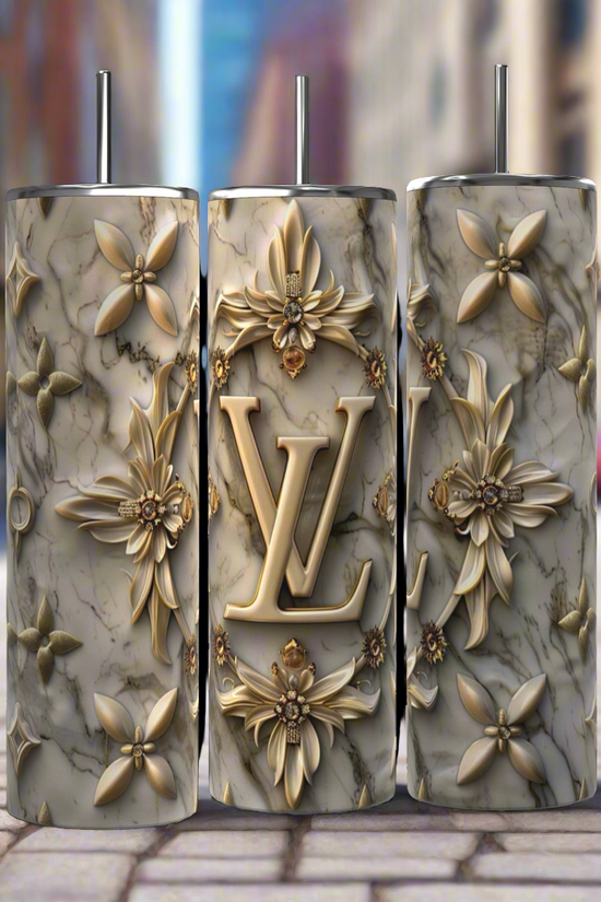 Three intricately decorated 20 oz Louis Vuitton Silver Gold tumblers by Kreative Kreationz, featuring a marbled texture and golden floral designs, stand side by side on a cobblestone street. The middle tumbler prominently showcases the initials 