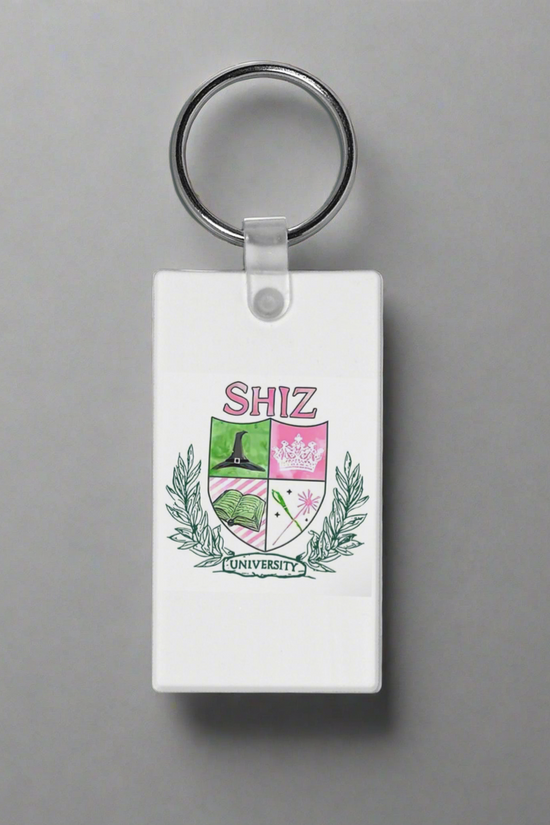 The Shitz University Keychain by Kreative Kreationz is a rectangular white accessory with a crest, green laurels, and shield icons—a book, castle, and pink flower—sublimation printed for vibrant detail.