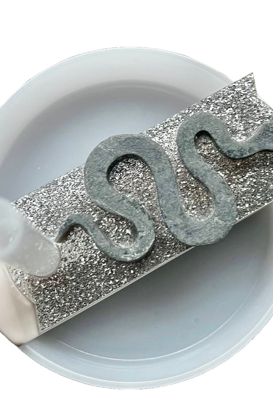 The REP 3D Snake Custom Stanley Plate/Topper by Kreative Kreationz features a coiled, snake-like shape made of a dark, ashy material on a rectangular, glittery silver surface. This setup, equipped with a high-quality acrylic lid topper, is placed inside a white circular container with a transparent tube on the left side.