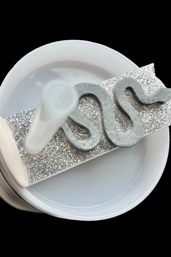 The REP 3D Snake Custom Stanley Plate/Topper by Kreative Kreationz features a coiled, snake-like shape made of a dark, ashy material on a rectangular, glittery silver surface. This setup, equipped with a high-quality acrylic lid topper, is placed inside a white circular container with a transparent tube on the left side.