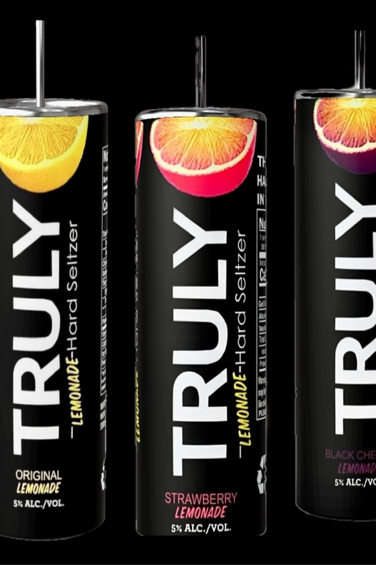 A lineup of Kreative Kreationz Truly 20oz Tumblers with add-on lid toppers is displayed on a sandy beach with a sunny, blurred background of the ocean and a distant island. The tumblers feature colorful fruit graphics reminiscent of Truly Flavors and come in stylish white and black designs, much like the trendy hard seltzer cans that keep beverages hot or cold.
