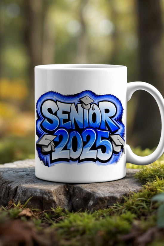 The Class Of 2025 Graduate Mug by Kreative Kreationz features blue graffiti-style text and graduation caps, displayed on a tree stump outdoors—an ideal graduation gift with a forest backdrop.