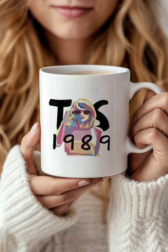 A person with long blonde hair and a white sweater holds the Taylor Swift 1989 Mug by Kreative Kreationz, featuring 
