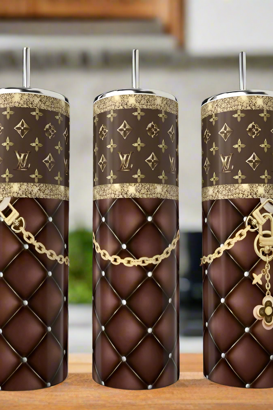 The Kreative Kreationz Louis Vuitton 20oz Tumblers feature brown quilted patterns, gold LV-inspired designs, and metal straws—luxury drinkware for any enthusiast. Three stainless steel tumblers are shown on a kitchen counter with a softly blurred background.