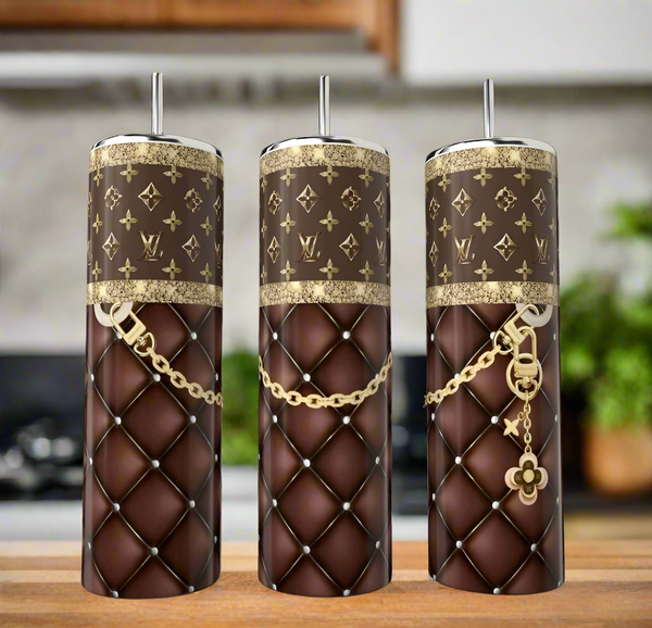 The Kreative Kreationz Louis Vuitton 20oz Tumblers feature brown quilted patterns, gold LV-inspired designs, and metal straws—luxury drinkware for any enthusiast. Three stainless steel tumblers are shown on a kitchen counter with a softly blurred background.