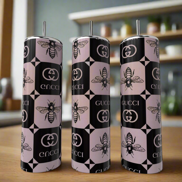 Three Gucci Pink & Black Inspired 20oz tumblers by Kreative Kreationz, featuring bee and logo motifs, sit on a wooden surface. Their luxury pattern in pink, black, and white stands out against a blurred kitchen background.