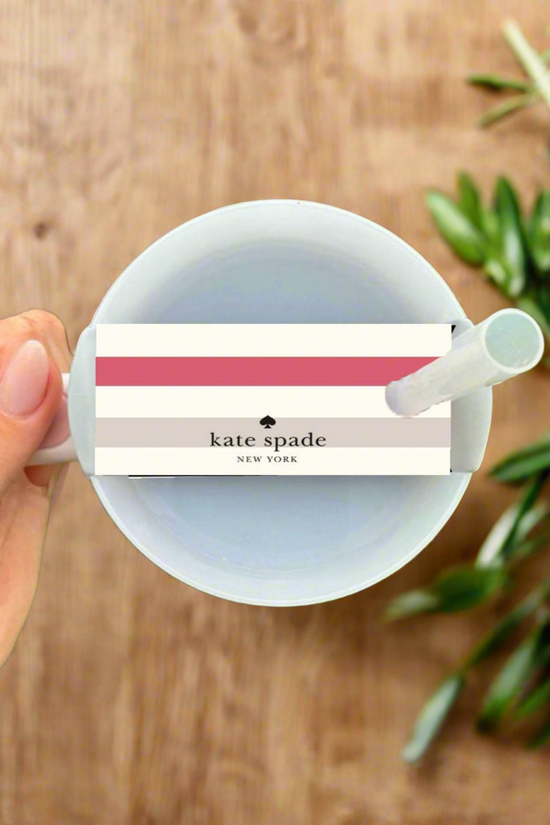 A hand holds a white cup topped with the Kate Spade Stripe Stanley Lid Topper by Kreative Kreationz, placed beside a green leafy plant on a wooden surface.