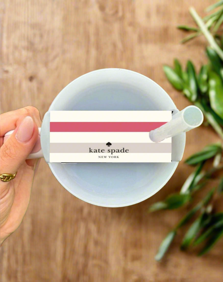 A hand holds a white cup topped with the Kate Spade Stripe Stanley Lid Topper by Kreative Kreationz, placed beside a green leafy plant on a wooden surface.