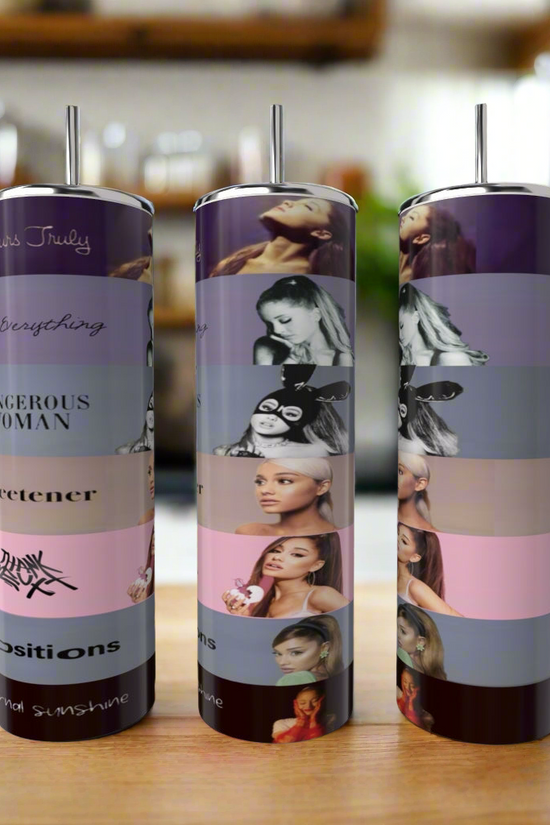 Three Kreative Kreationz Ariana Grande 20oz tumblers with metal straws sit on a kitchen counter, each decorated in vibrant horizontal bands featuring artistic portraits, song titles, and album covers.