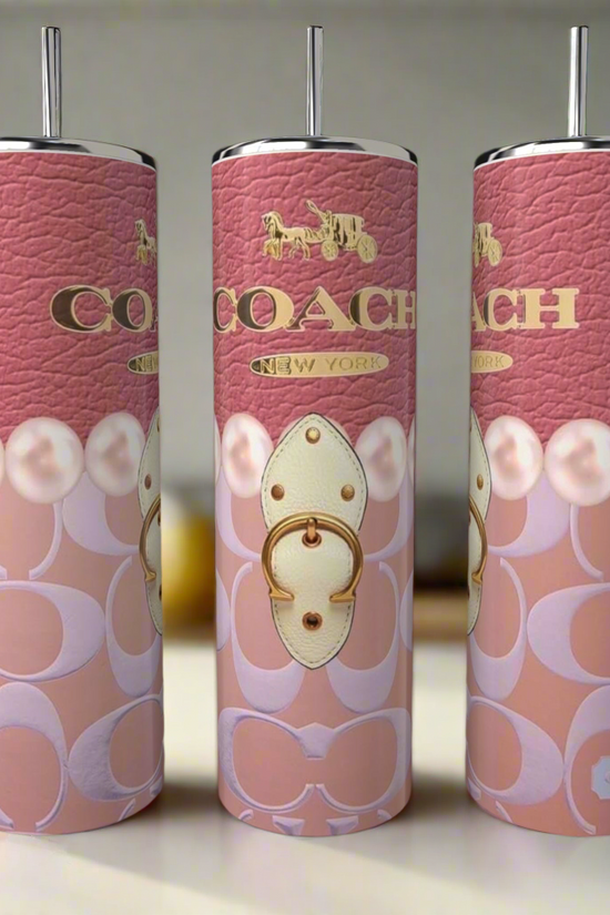 Three pink Kreative Kreationz Coach 20oz Skinny Tumblers, complete with metal straws, feature the luxury-inspired Coach logo, a gold horse and carriage motif, and intricate patterns on the counter, while kitchen items appear blurred in the background.