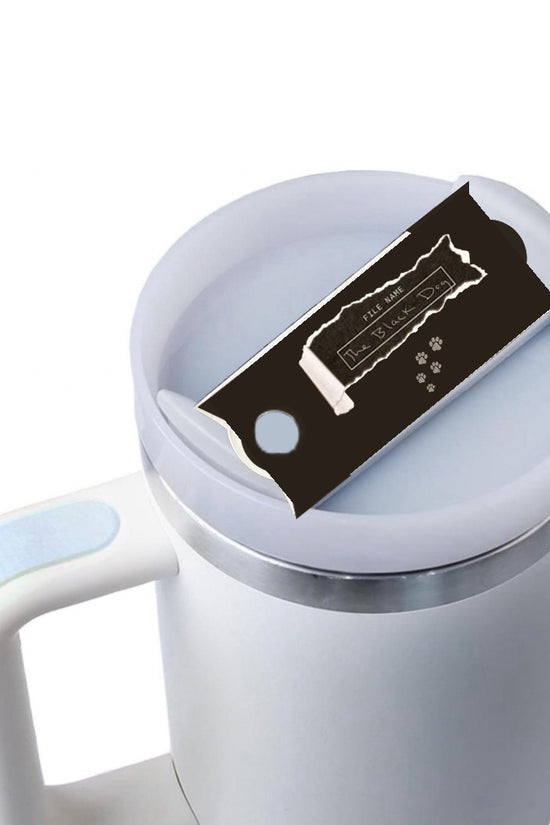 A white mug with a light blue handle is shown against a plain background. On top of the Kreative Kreationz Black Dog lid Plate/Topper, which fits 20, 30, and 40 oz Stanley cups, is a partially unwrapped chocolate bar with a black wrapper.