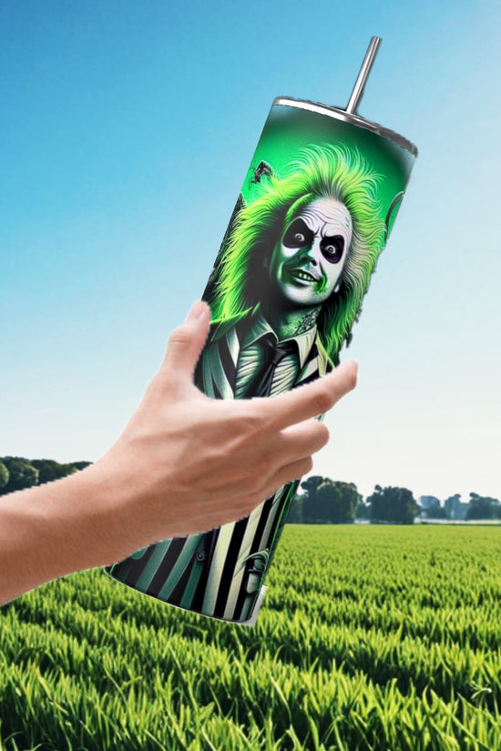 A hand holds the Beetlejuice Neon 20oz Tumbler by Kreative Kreationz, showcasing a vibrant design of a green-haired character in a striped suit. Set against a lush green field and blue sky, this drinkware is ideal for Halloween sips.