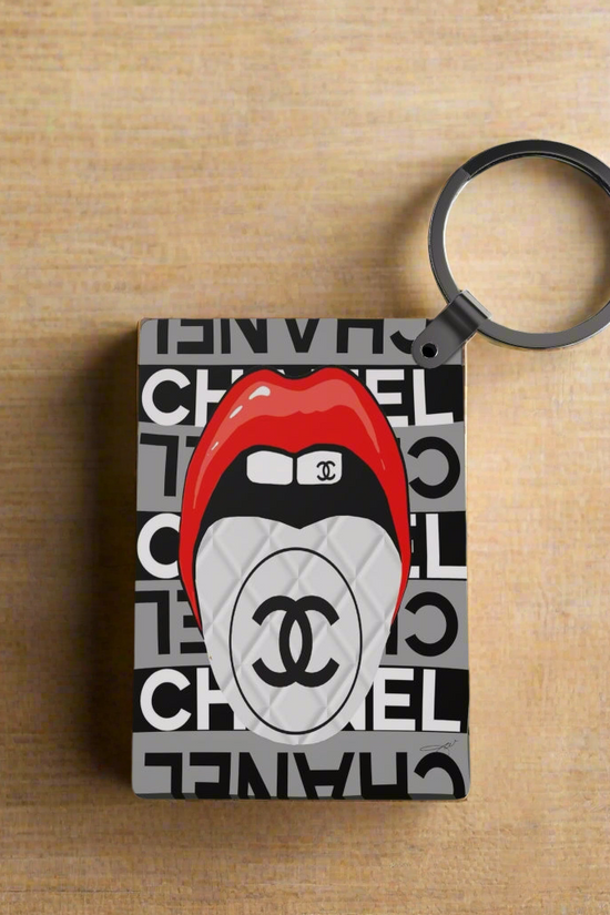 The Designer Inspired Keychain by Kreative Kreationz features a rectangular design with a bold Chanel-style logo, red lips biting a white double C emblem, all set against repeated black and white Chanel-inspired text.