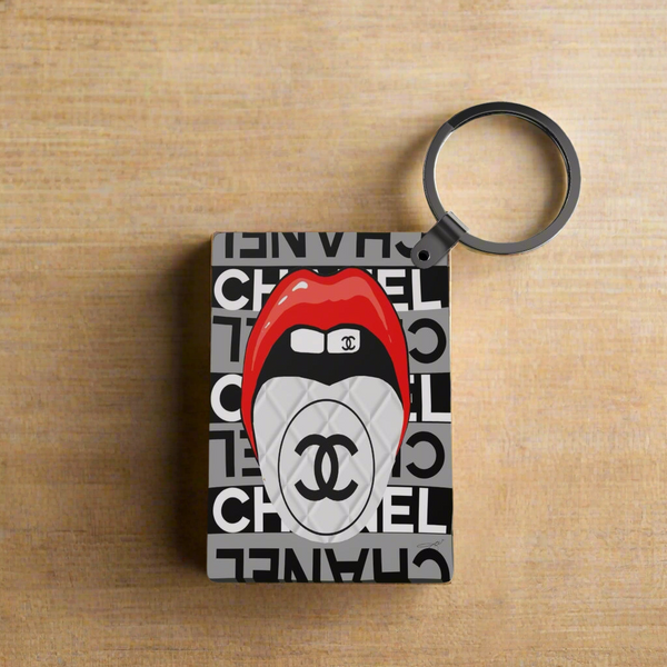 The Designer Inspired Keychain by Kreative Kreationz features a rectangular design with a bold Chanel-style logo, red lips biting a white double C emblem, all set against repeated black and white Chanel-inspired text.