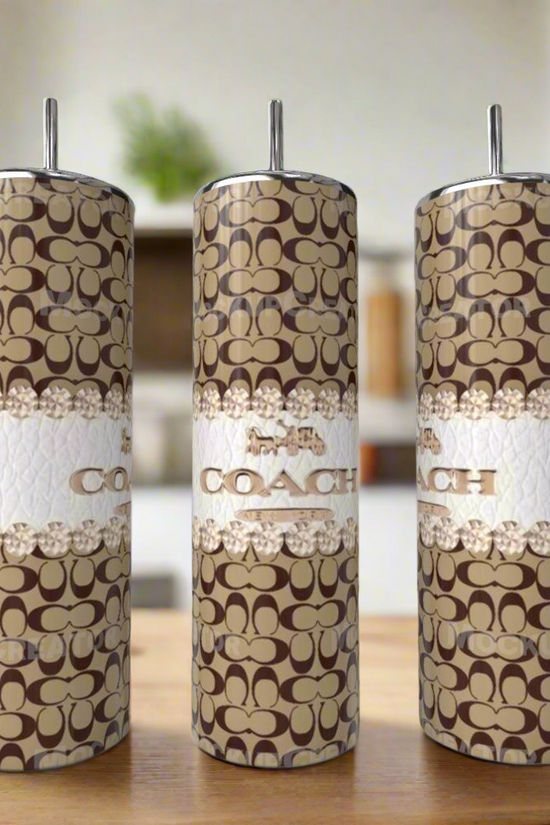Three Kreative Kreationz Coach Brown 20oz Skinny Tumblers, featuring brown and beige logo patterns with white bands, sit on a wooden surface, adding stylish luxury to the modern kitchen.