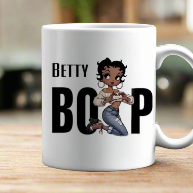 The Betty Boop Ceramic Mug by Kreative Kreationz is a white 11oz ceramic mug with "Betty Boop" text and a cartoon of Betty Boop kneeling, set on a blurred indoor background. Dishwasher safe.